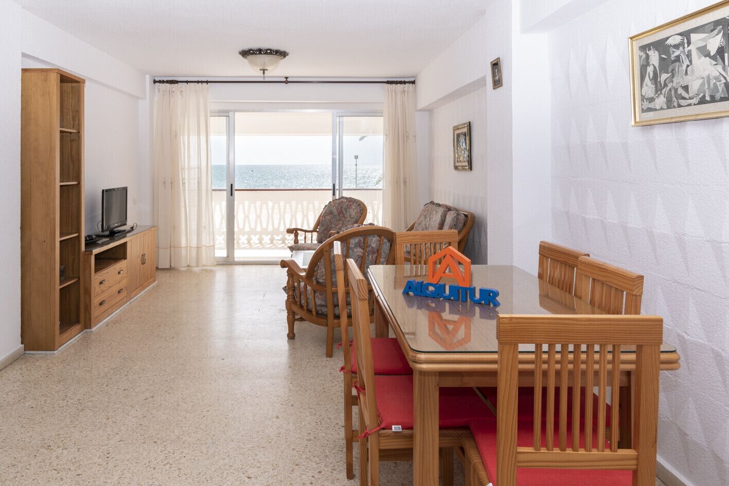 A spacious room with a wooden dining table, chairs, a TV on a wooden cabinet, and a balcony with a view of the sea.