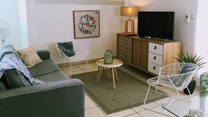 Un salon moderne avec un canapé gris, un fauteuil blanc, une petite table avec une lampe, un téléviseur posé sur un meuble en bois et une œuvre d’art encadrée accrochée au mur.