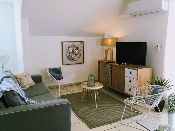 A modern living room with a grey sofa, a white chair, a small table with a lamp, a TV on a wooden cabinet, and a framed artwork on the wall. - Grand T3 charming 6min walk from the lagoon (La Saline-Les-Bains)