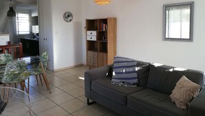 Un salon moderne avec un canapé gris, une bibliothèque, un coin repas avec des chaises et, au fond, une cuisine.