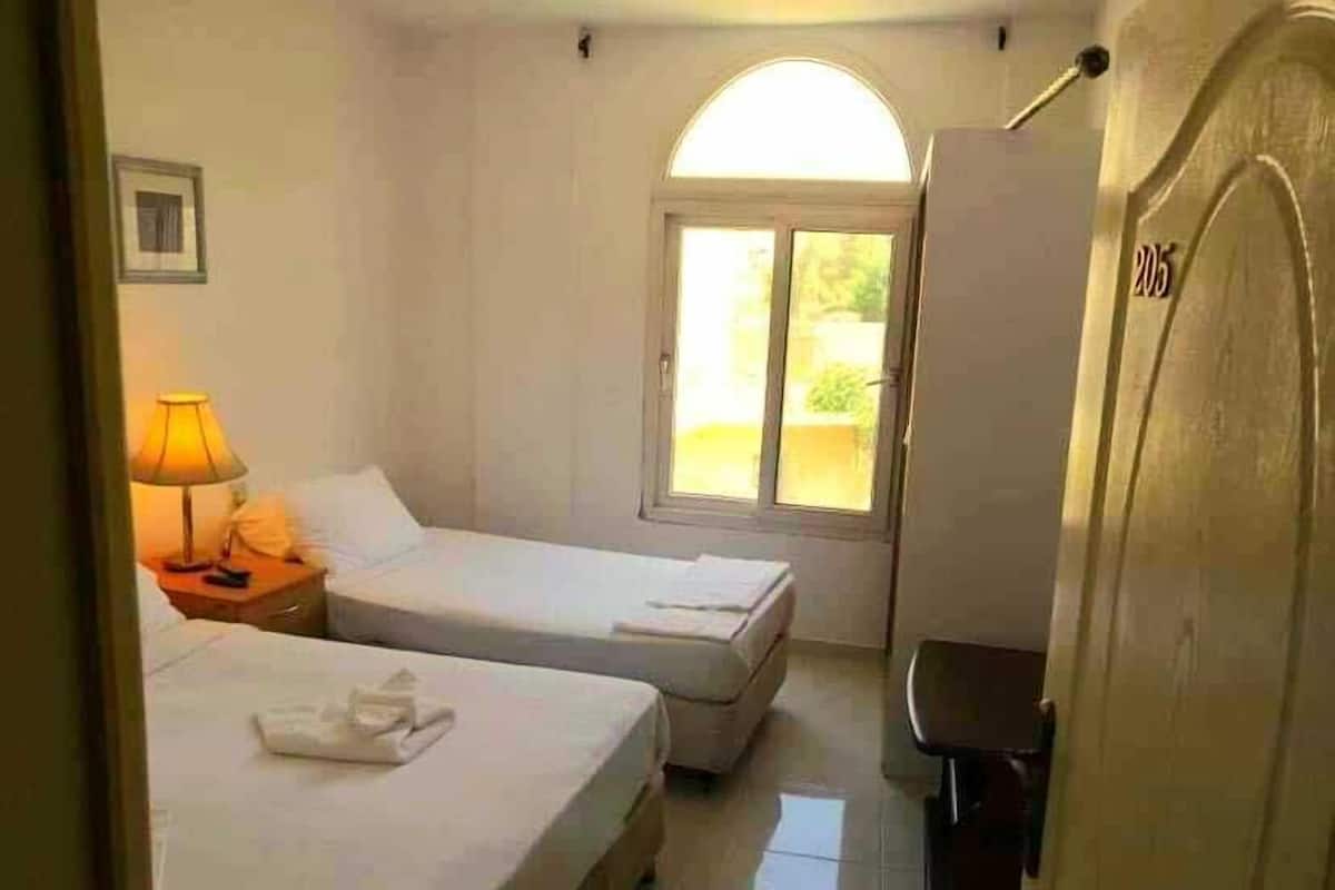 Une chambre d’hôtel avec deux lits, une table de chevet avec une lampe, une fenêtre donnant sur un paysage agréable et une porte portant un numéro.