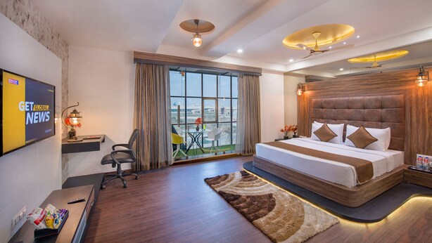A modern hotel room with a large bed, a desk with a computer, a TV displaying news, and a balcony with a city view.