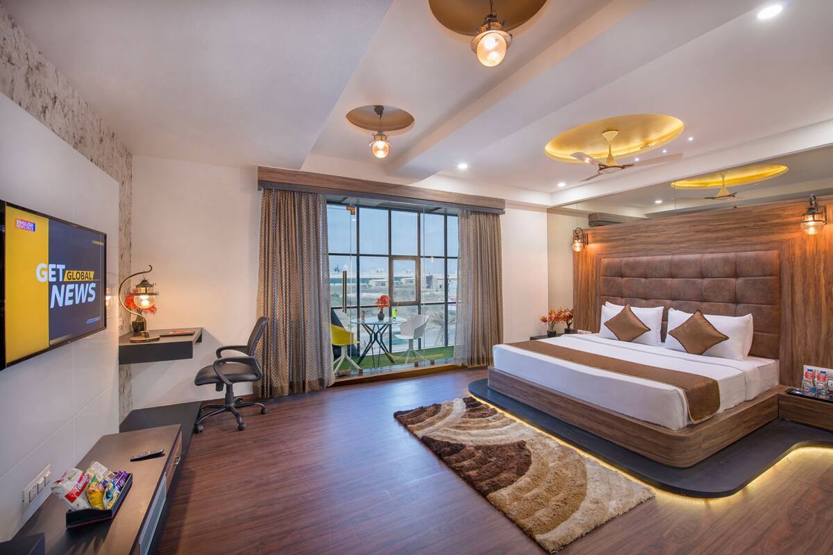 Quarto de hotel moderno com uma cama grande, uma escrivaninha com computador, uma televisão exibindo notícias e uma varanda com vista para a cidade.