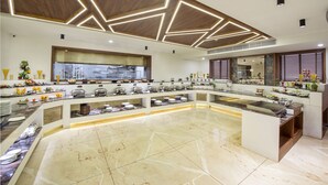 A modern hotel lobby with a central island featuring a buffet, a wooden ceiling design, and a large window providing a view of the kitchen.
