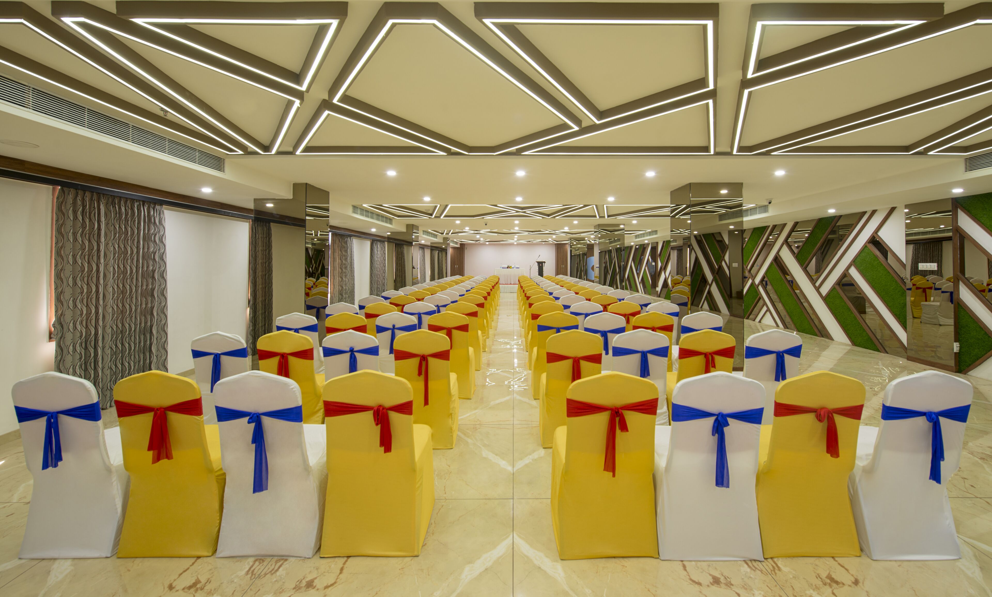 A conference room with rows of chairs, each with a white and yellow cover and red and blue sashes. The room has a modern ceiling design with geometric patterns and recessed lighting.