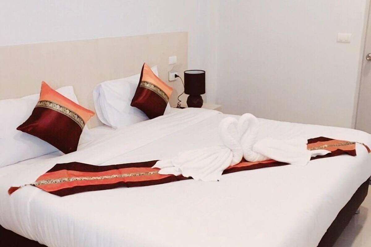 A neatly made bed with white linens and decorative pillows, a bedside table with a lamp, and a plain wall.