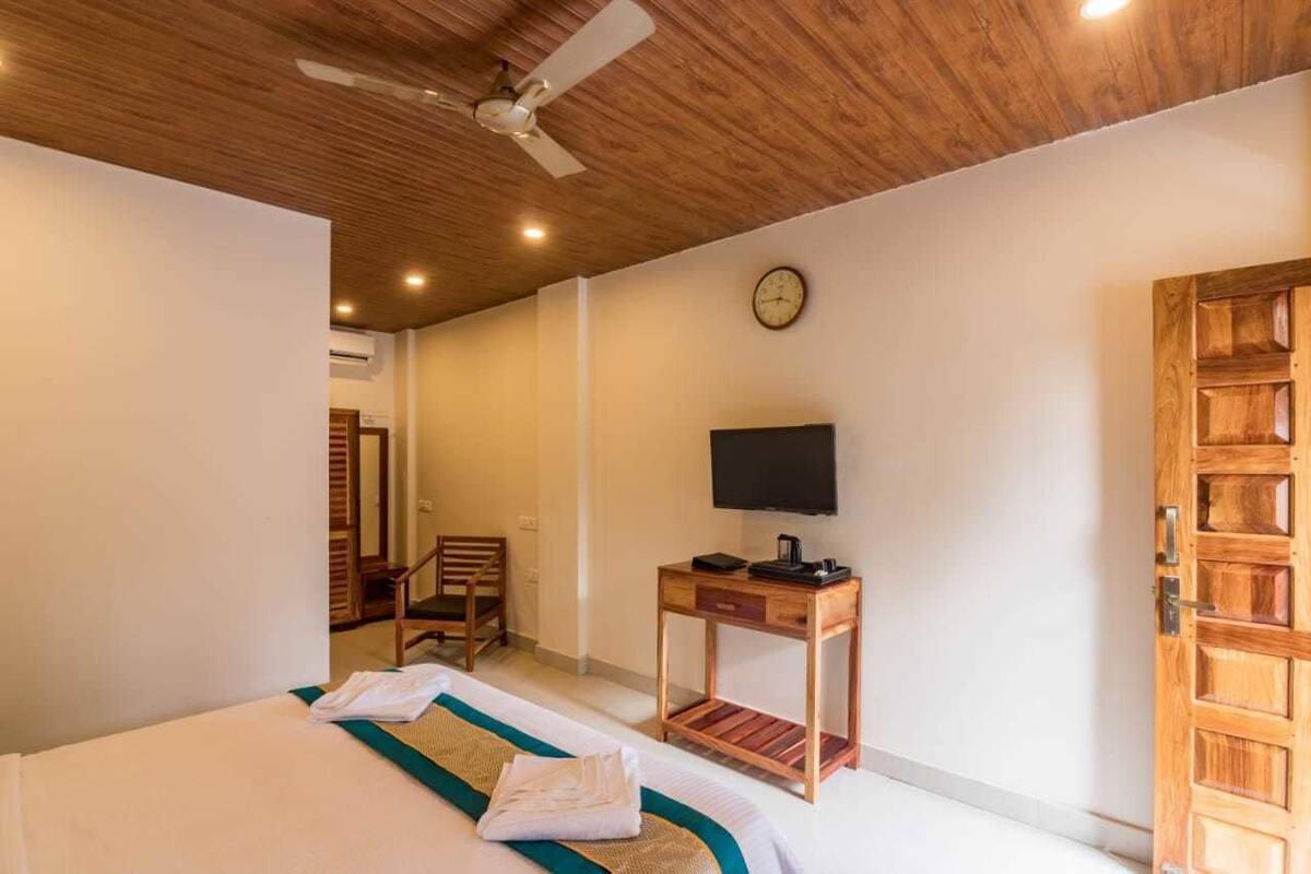 A hotel room with a wooden ceiling, a bed with a green and white bedspread, a wooden nightstand with a TV, a wooden door, and a chair.