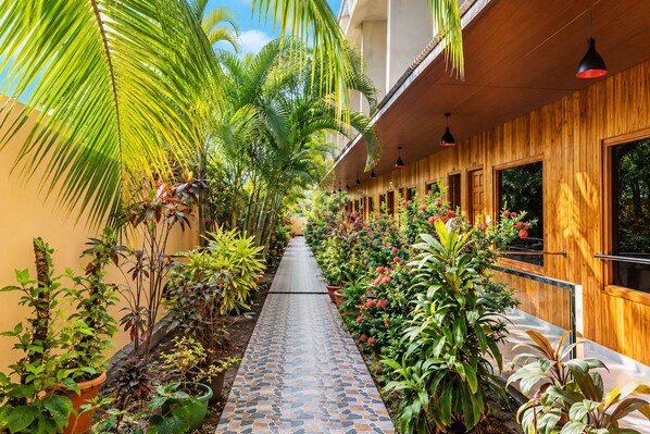 A corridor with a tiled floor, surrounded by various plants and a wooden building with windows.