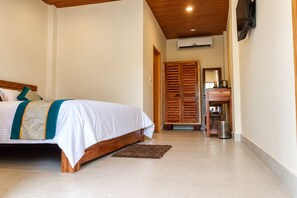 A bedroom with a bed, a wooden wardrobe, a desk with a chair, and a trash bin.