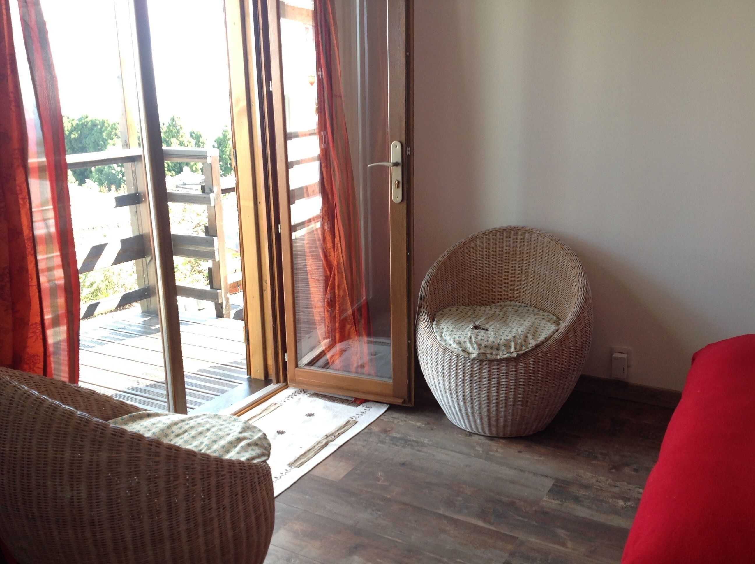 Ein Zimmer mit einer Schiebetür zum Außenbereich, ein Korbstuhl mit Sitzkissen und ein rotes Sofa.
