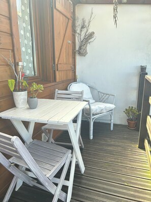 Um deck de madeira com uma mesa e cadeiras brancas, plantas em vasos e uma parede rústica.