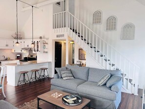 Ein geräumiger Wohnbereich mit grauem Sofa, Holztisch und einer Küche mit weißen Schränken und einer weißen Treppe.