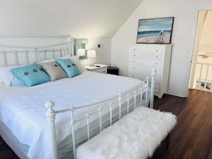 Un dormitorio con una cama blanca, un banco blanco, una mesita de noche, un aparador y un cuadro en la pared.