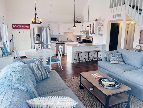 Una sala de estar moderna con sofá, mesa de centro y área de cocina con armarios blancos y electrodomésticos de acero inoxidable.