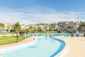 Una piscina con bordo curvo, circondata da una zona pavimentata con palme e una fila di edifici colorati sullo sfondo.