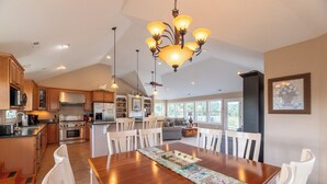 A spacious kitchen with modern appliances, a dining area with a large wooden table and white chairs, and a chandelier. - Show Boat - Kitty Hawk MP 2.5, Pool, Hot Tub, Rec Room, Pool Table, Golf Course Views (Kitty Hawk)