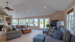 A spacious living room with a fireplace, a ceiling fan, and a large window overlooking a deck. - Show Boat - Kitty Hawk MP 2.5, Pool, Hot Tub, Rec Room, Pool Table, Golf Course Views (Kitty Hawk)