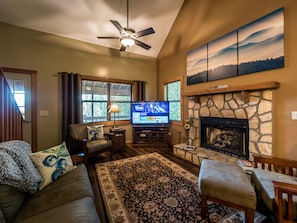 A cozy living room with a stone fireplace, a ceiling fan, and a television displaying a movie. - 🍁 Huckleberry Cabin - wifi, Fireplace,  fire pit, VIEWS! Only 3 miles To Helen! (Cleveland)
