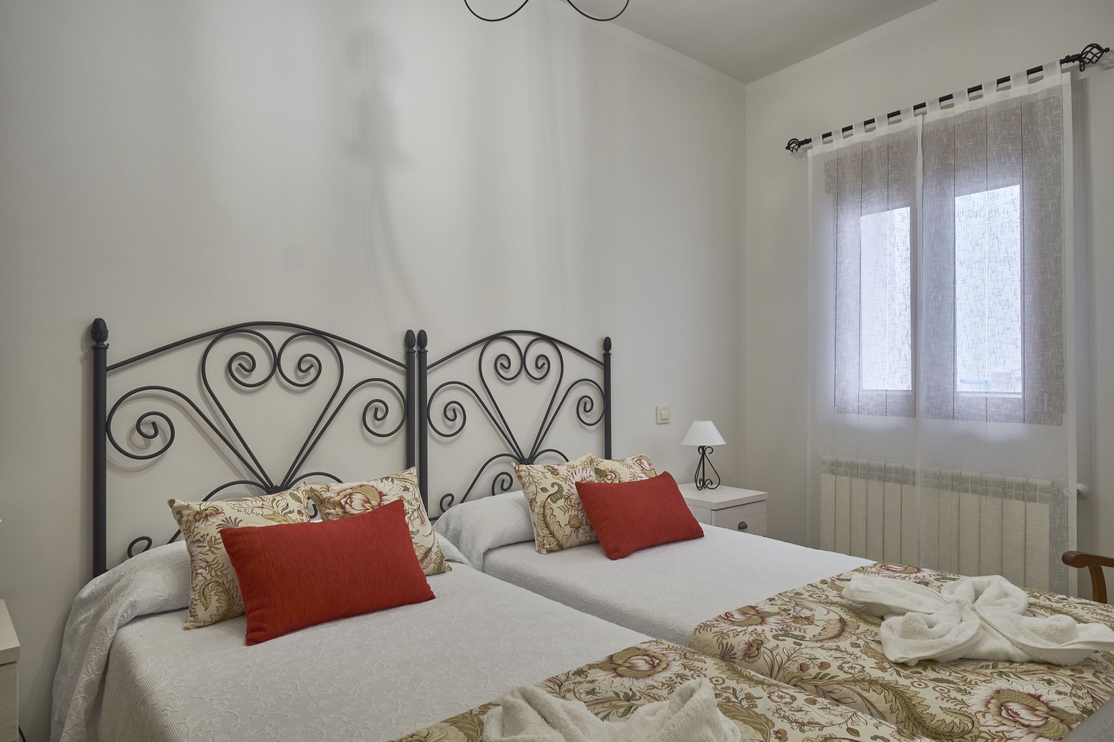 A bedroom with two beds, each with patterned bedding and red pillows. There is a window with sheer curtains, a bedside table with a lamp, and a radiator.