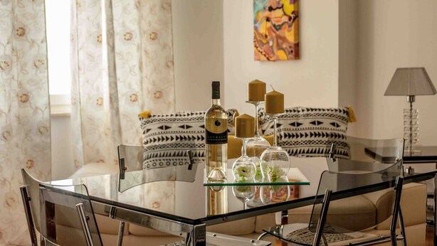 Ein modernes Wohnzimmer mit einem Glastisch, einer Weinflasche, Kerzen und dekorativen Blumen. Es gibt ein Sofa mit gemusterten Kissen und ein Bild an der Wand.