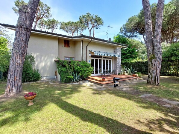 A house with a covered patio, a barbecue grill, and a garden with trees and a flower pot.