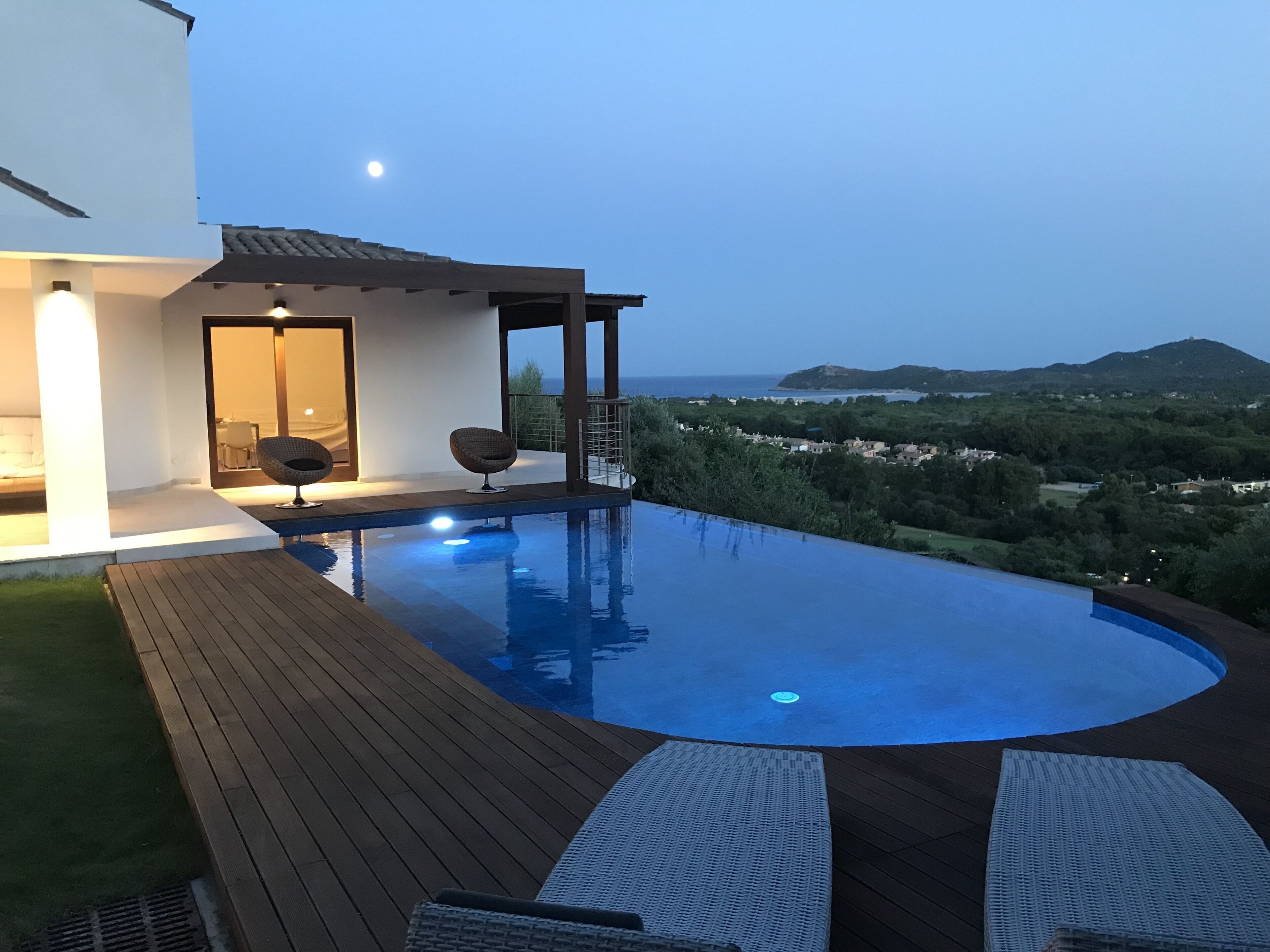 Ein moderner Infinity-Pool mit Holzterrasse, Liegestühlen, einer kleinen überdachten Sitzgelegenheit und Blick auf die Landschaft.