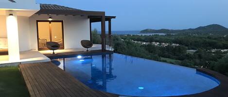 Ein moderner Infinity-Pool mit Holzterrasse, Liegestühlen, einer kleinen überdachten Sitzgelegenheit und Blick auf die Landschaft.