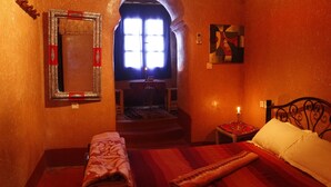 A bedroom with a bed, a nightstand, a lamp, a window, and a painting on the wall. - Guest House The Dor fibula (Aït Ben Haddou)