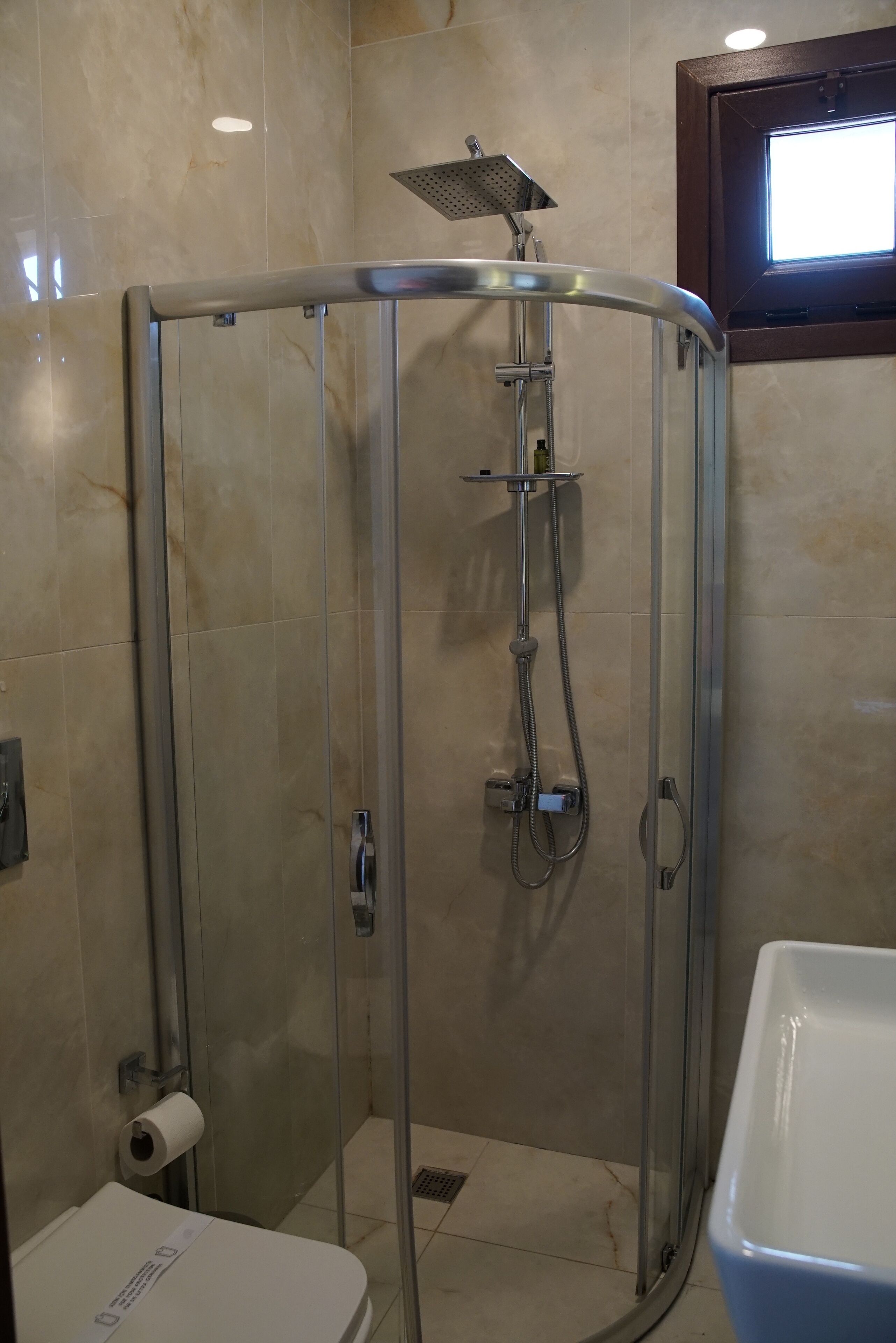 Cabina de ducha con paredes de vidrio, cabezal de ducha manual, inodoro con dispensador de toallas de papel y lavamanos blanco.