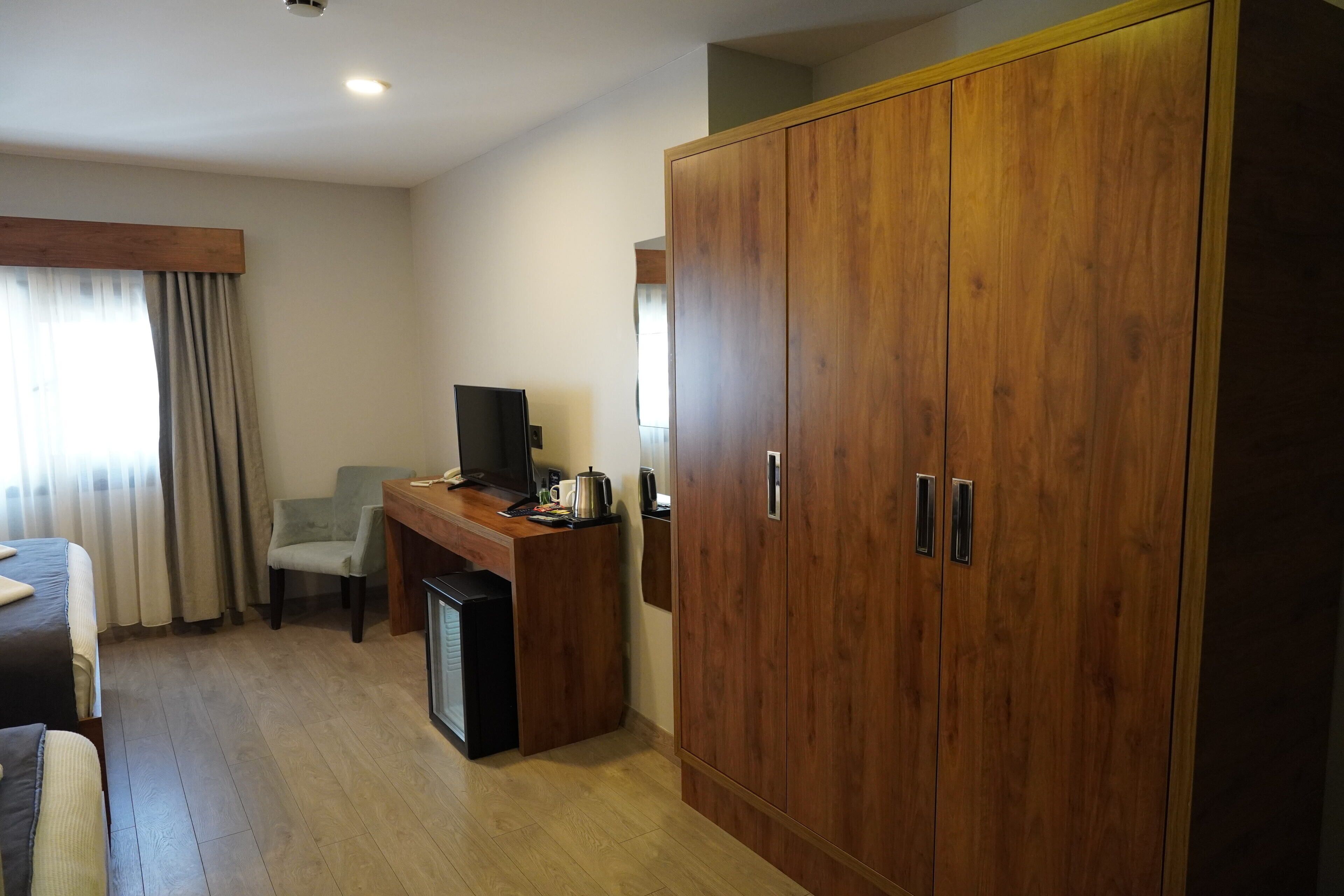 A hotel room with a wooden wardrobe, a desk with a television, a chair, and a bed.