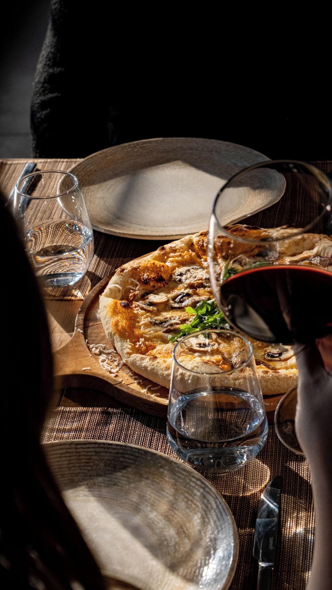 A pizza on a wooden board with a glass of red wine and a glass of water on a table.