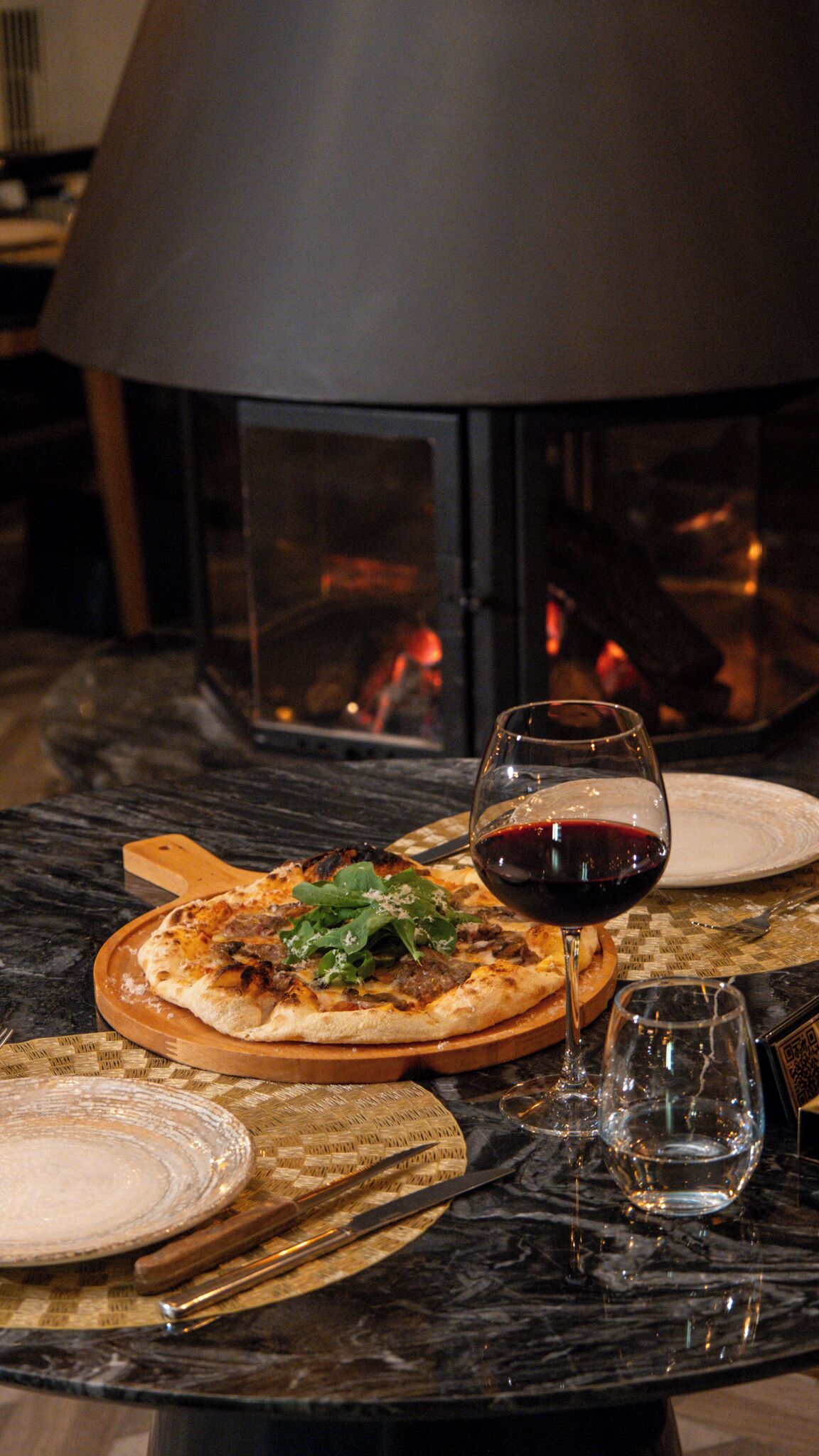 A pizza on a wooden board, a glass of red wine, and a table setting with a knife and spoon.