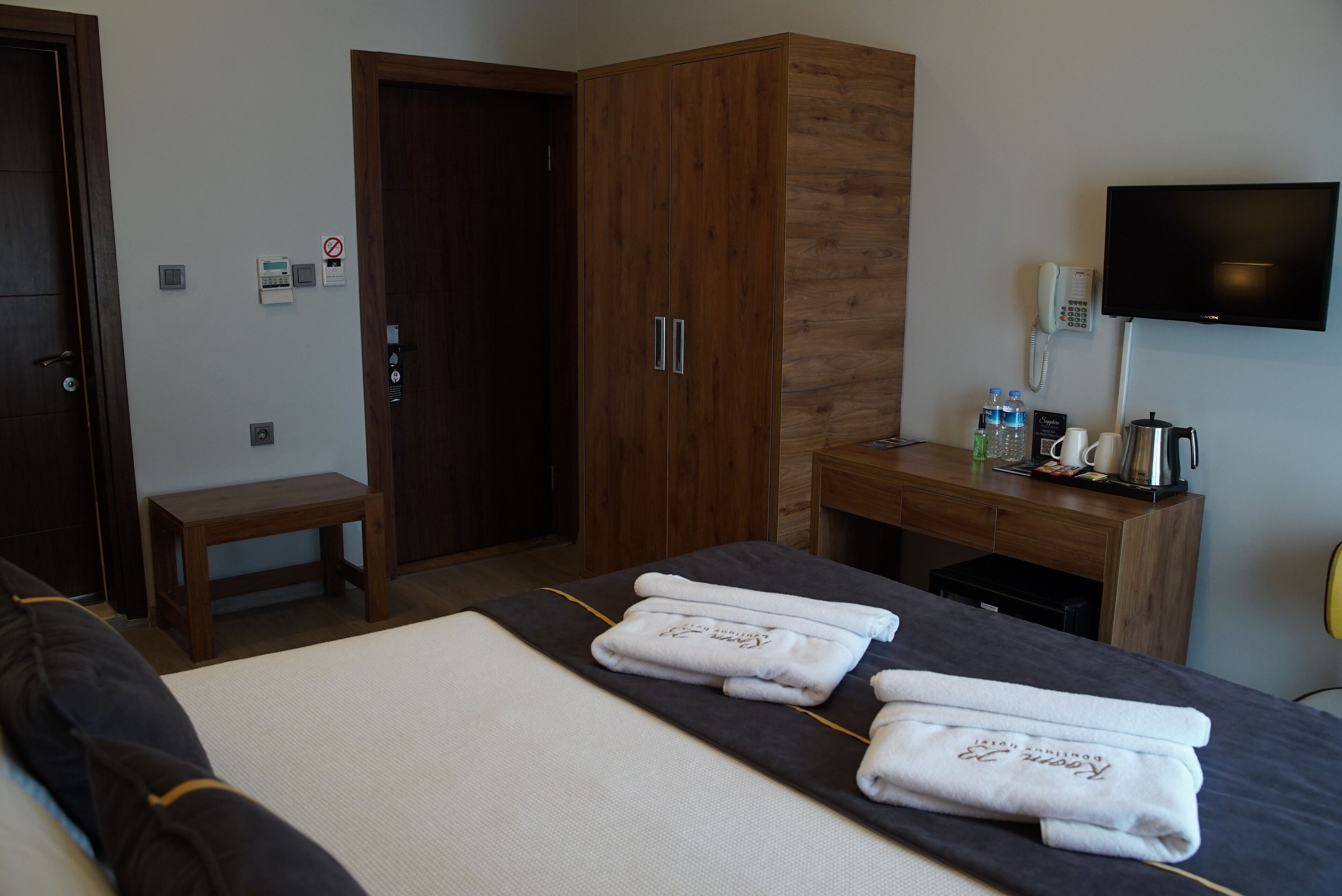 Habitación de hotel con una cama, un armario de madera, un escritorio con teléfono y botellas de agua, y un televisor en la pared.