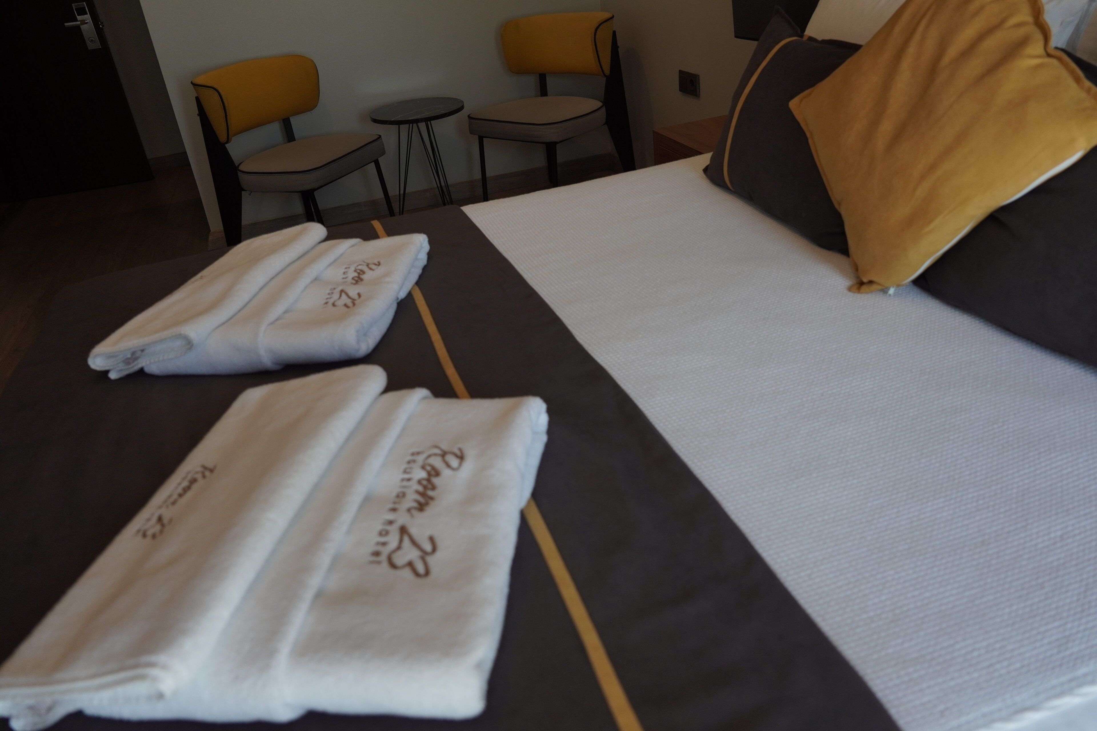 A hotel room with a neatly made bed, two towels with embroidered text, a yellow pillow, and two chairs.