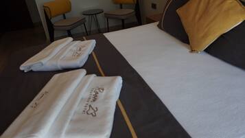 A hotel room with a neatly made bed, two towels with embroidered text, a yellow pillow, and two chairs.