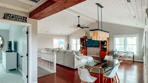 A spacious living area with a glass dining table, white chairs, a ceiling fan, and a kitchen area with a TV and bookshelves.
