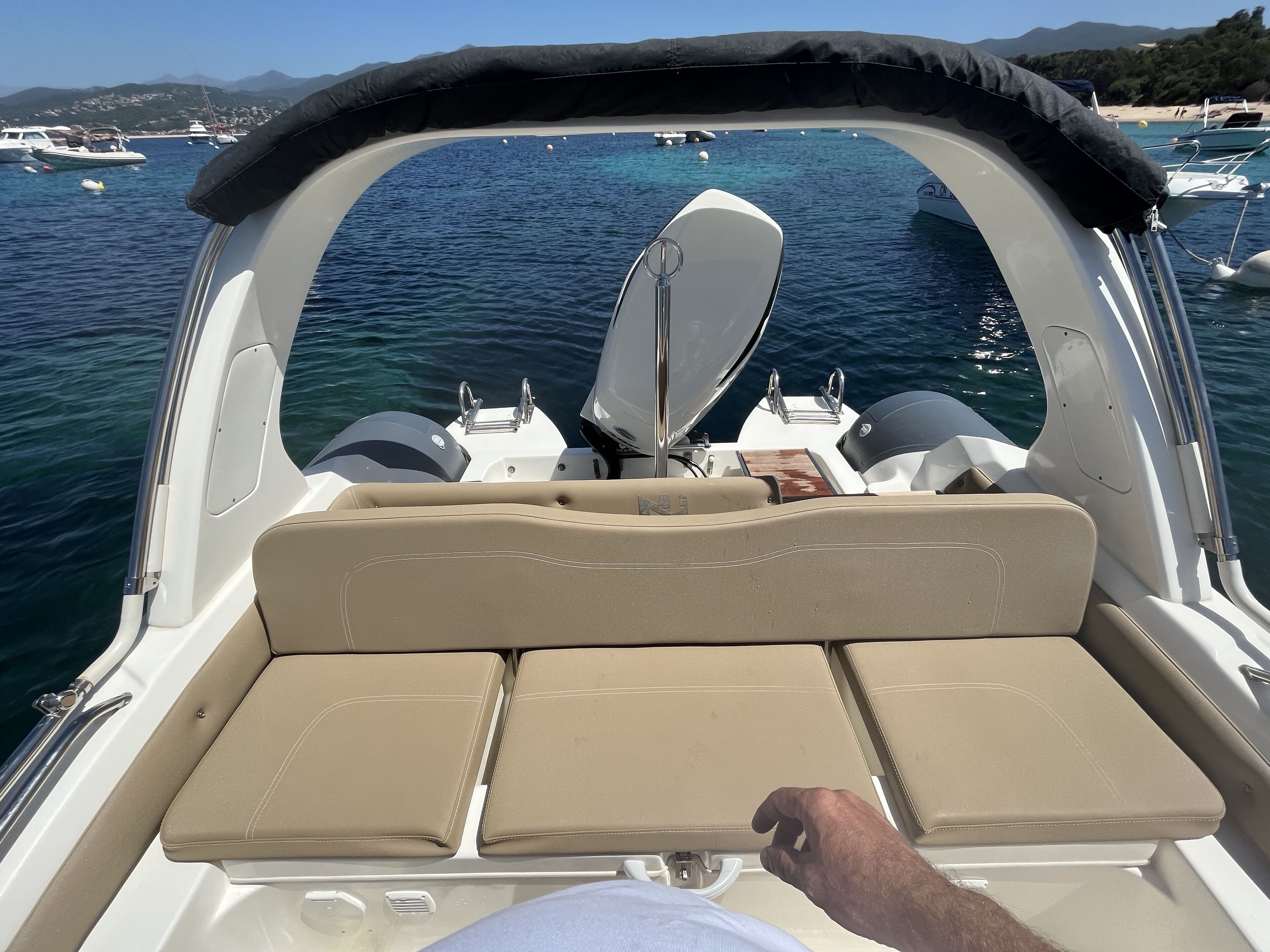 A boat's interior with a cushioned seating area, a steering wheel, and a view of the water with other boats and a beach in the background.