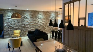 A modern living room with a stone wall, a yellow armchair, a dark sofa, and a wooden dining table with hanging pendant lights.