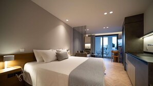 A modern hotel room with a large bed, bedside table, and a view of the city. - Alinea Suites Limassol Center (Limassol)