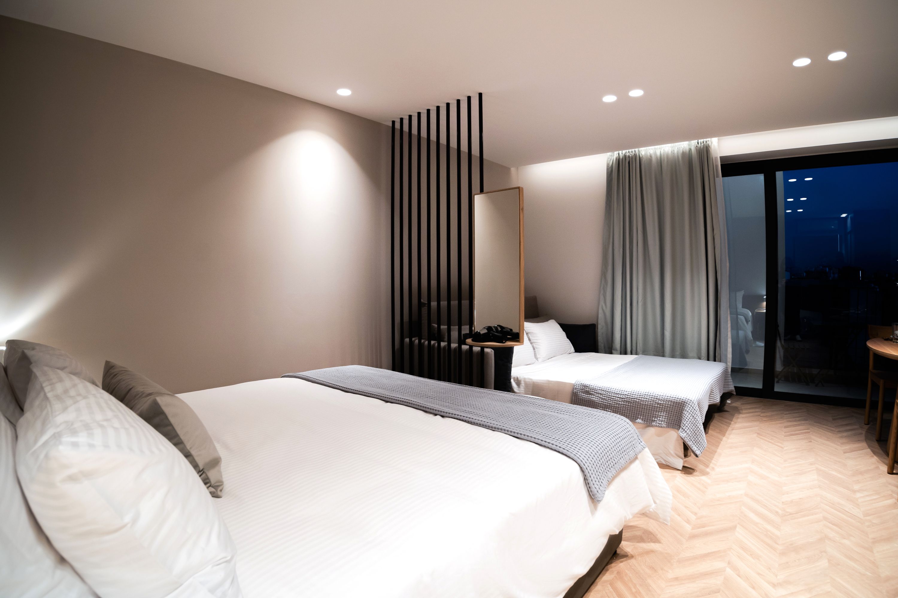 A modern hotel room with two beds, a large mirror, a desk, and a view of the city at night.