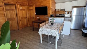 Ein gemütliches Blockhauszimmer mit Holzwänden, einem Esstisch mit Blumenmuster bedeckter Tischdecke, einem Fernseher, einer Küchenzeile inklusive Kühlschrank und einer großen Pflanze.