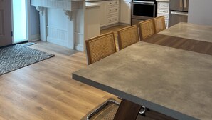 A modern kitchen with white cabinetry, a stainless steel refrigerator, a microwave, and an oven. There is a dining area with a long table and chairs.