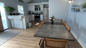 A modern kitchen with white cabinetry, a stainless steel refrigerator, and a dining area with a long table and wicker chairs.