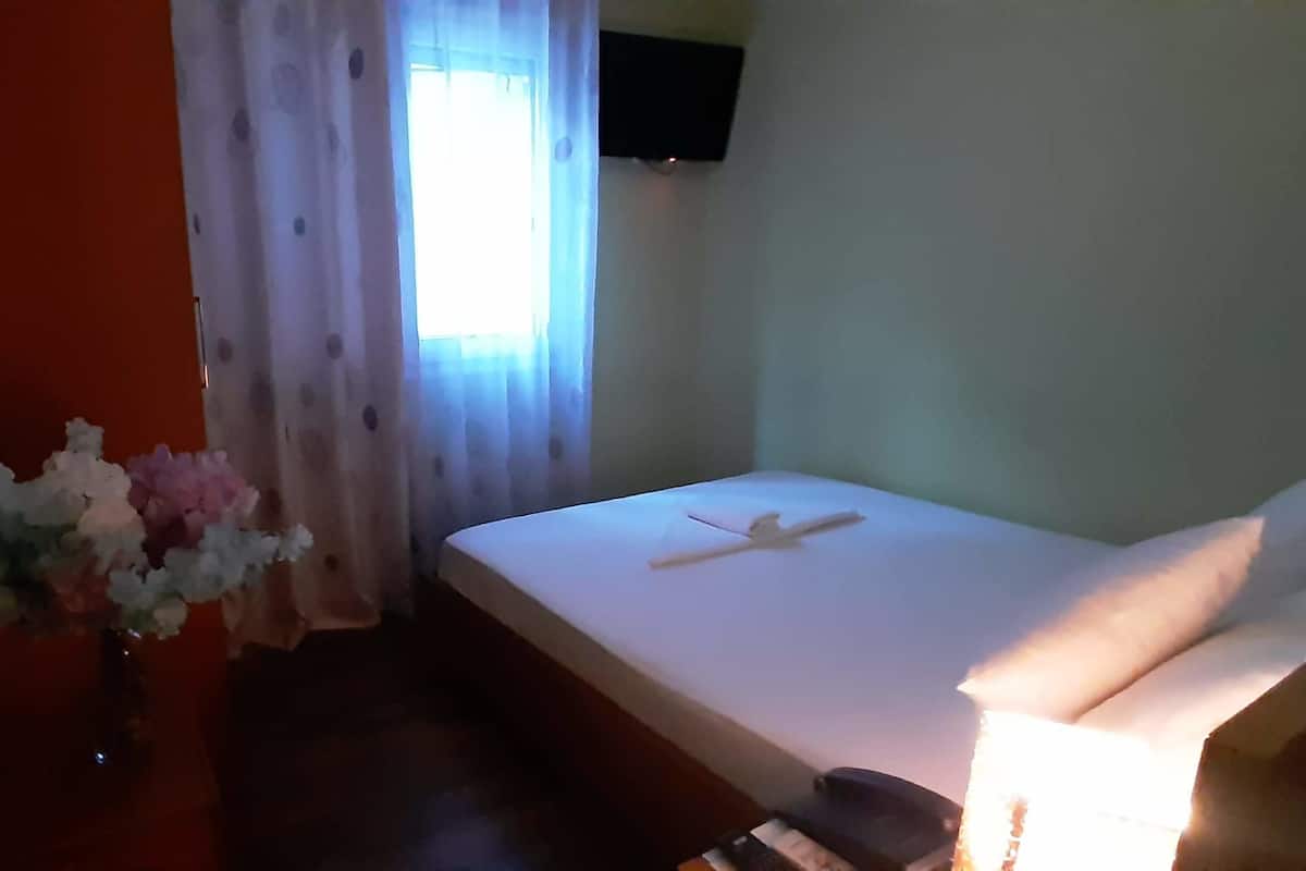 Ein Hotelzimmer mit einem Bett, einem kleinen Tisch mit Blumen in einer Vase, einem Fenster mit Vorhängen und einem flachbildfernseher an der Wand.