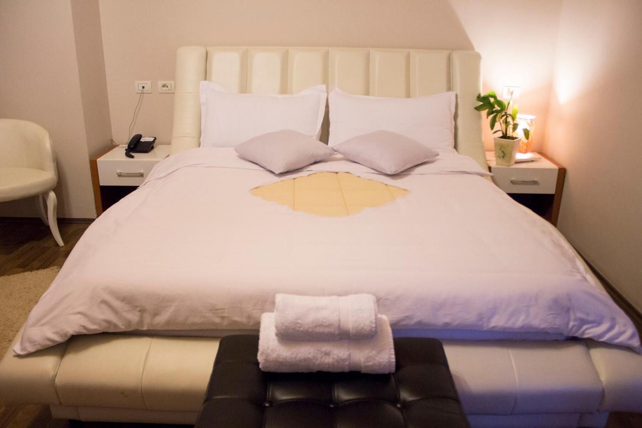 Una cama bien hecha, con un cabecero blanco, dos almohadas y una toalla doblada sobre un banco. Se ve una mesita de noche con una lámpara y un teléfono.