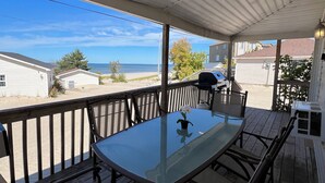Een overdekt terras buiten met een glazen tafel en stoelen, met uitzicht op een strand en woongebouwen.