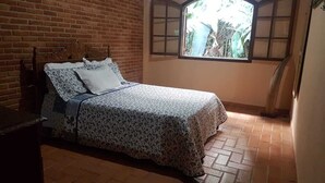 A bedroom with a bed, a window with a view of greenery, and a brick wall. - HOUSE IN PENEDO WITH WATERFALL (Penedo)