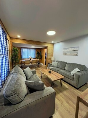 A modern living room with a grey sofa, a wooden coffee table, and a dining area with a table and chairs. - BEACHFRONT COTTAGE IN WASAGA BEACH - SANDPIPER BEACH RESORT - COTTAGE UNIT # 12 (Wasaga Beach)