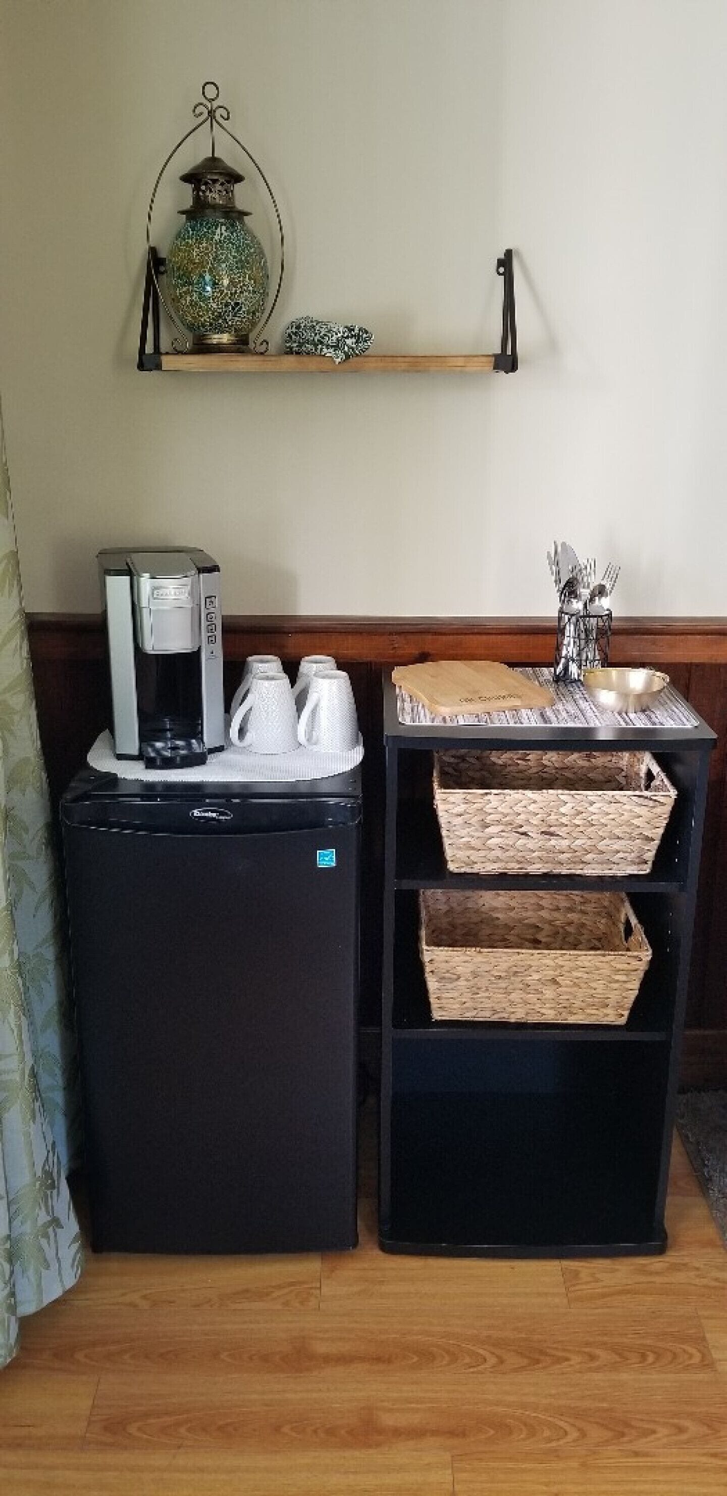 A compact kitchenette with a coffee maker, two white mugs, and a small shelf with a decorative item and a metal container.