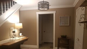 Un pasillo con una escalera, una lámpara, una mesita con una planta y una puerta que da a otra habitación.